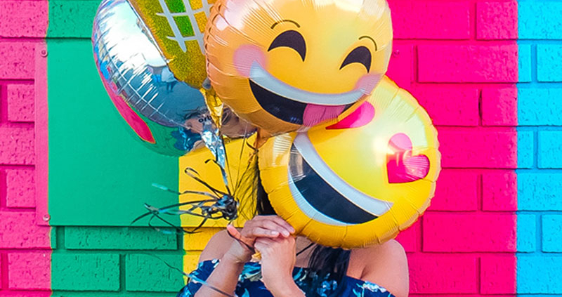 Woman standing in front of colorful wall holding smiley face balloon over her face